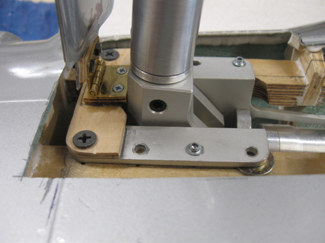 Tight shot of the outer door hinge during preliminary assembly. By butting the brass hinge up to the base of the retract, you can be assured of being parallel and in same plane as the axis pin for the retract. The smaller plywood piece under the hinge is a height adjustment for the door to be level with the bottom of the wing. The larger plywood cross piece serves as a mount for the hinge as there is nothing there to begin with. Also note the plywood “wedge” under the upper half of the hinge to offset the door with the slope of the wing toward the leading edge. Note the wide cutout around the retract base. This area is closed off with lithoplate sheet later. 